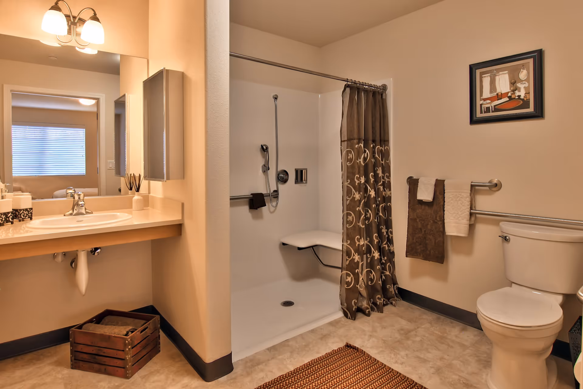 A clean and accessible bathroom featuring a walk-in shower with a fold-down seat and a decorative brown shower curtain. There is a white toilet with grab bars on the wall next to it, and two towels hanging on the bars. A large mirror is above a beige countertop with a sink, and a wooden crate with towels is placed on the floor beneath the sink. The bathroom has beige walls and a framed picture hanging above the toilet.