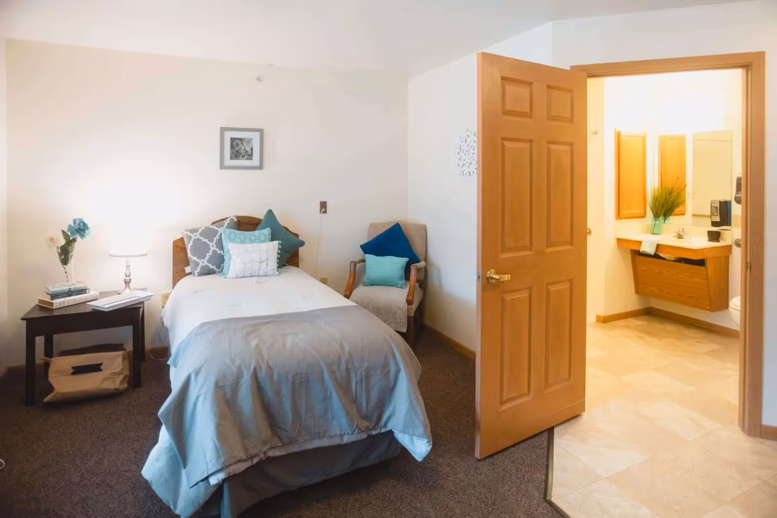A cozy senior living bedroom with a single bed made with light blue and gray bedding and multiple decorative pillows. Next to the bed is a small dark wooden nightstand with a lamp, books, and a vase with flowers. There is a cushioned armchair with two pillows in the corner. An open wooden door leads to a bathroom with a sink, mirror, and a plant on the counter.
