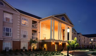 Exterior view of a multi-story senior living facility building at dusk with warm lights illuminating the entrance supported by large white columns, landscaped greenery, and a street lamp in front.