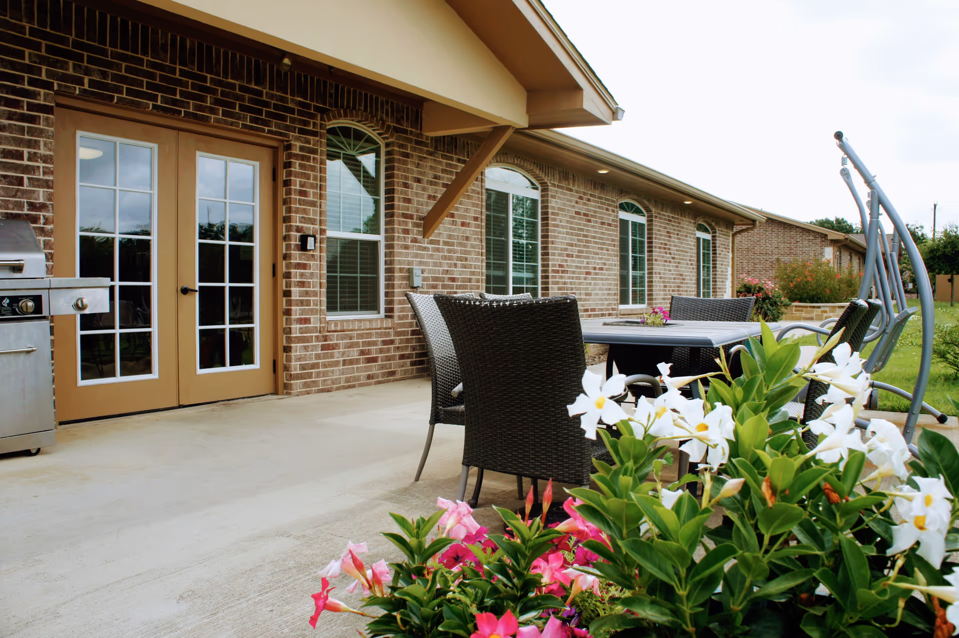Outdoor patio area at Duncanville Senior Living with a brick exterior wall, glass double doors, several windows, a stainless steel grill, black wicker chairs around a table, a swing, and colorful flowers in the foreground.