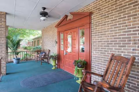 Brick building entrance with red door, porch seating, and potted plants on a covered outdoor walkway.