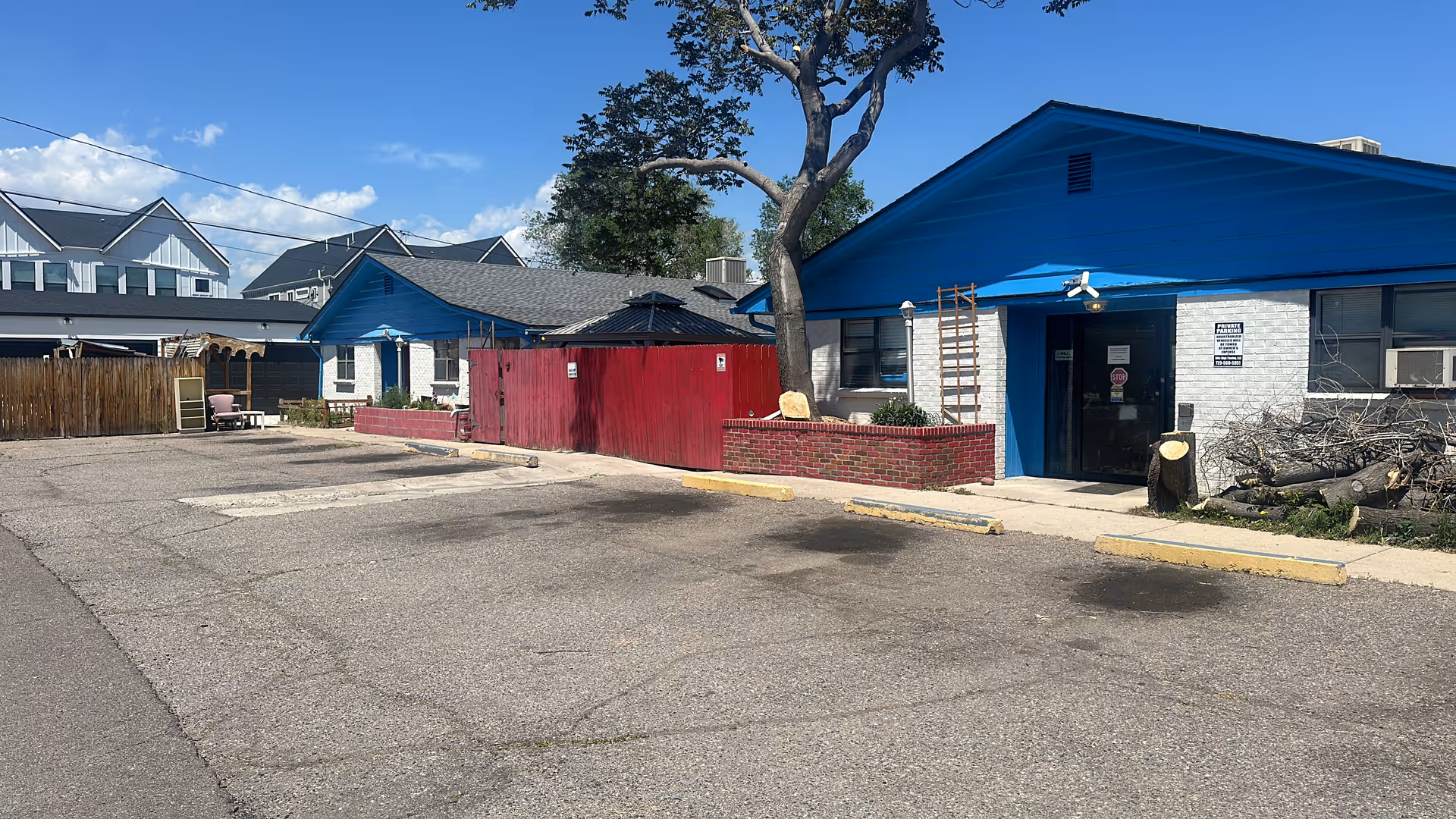 Exterior view of a single-story building with a blue roof and white brick walls, a red wooden fence, a tree, and a paved parking area in front under a clear blue sky.