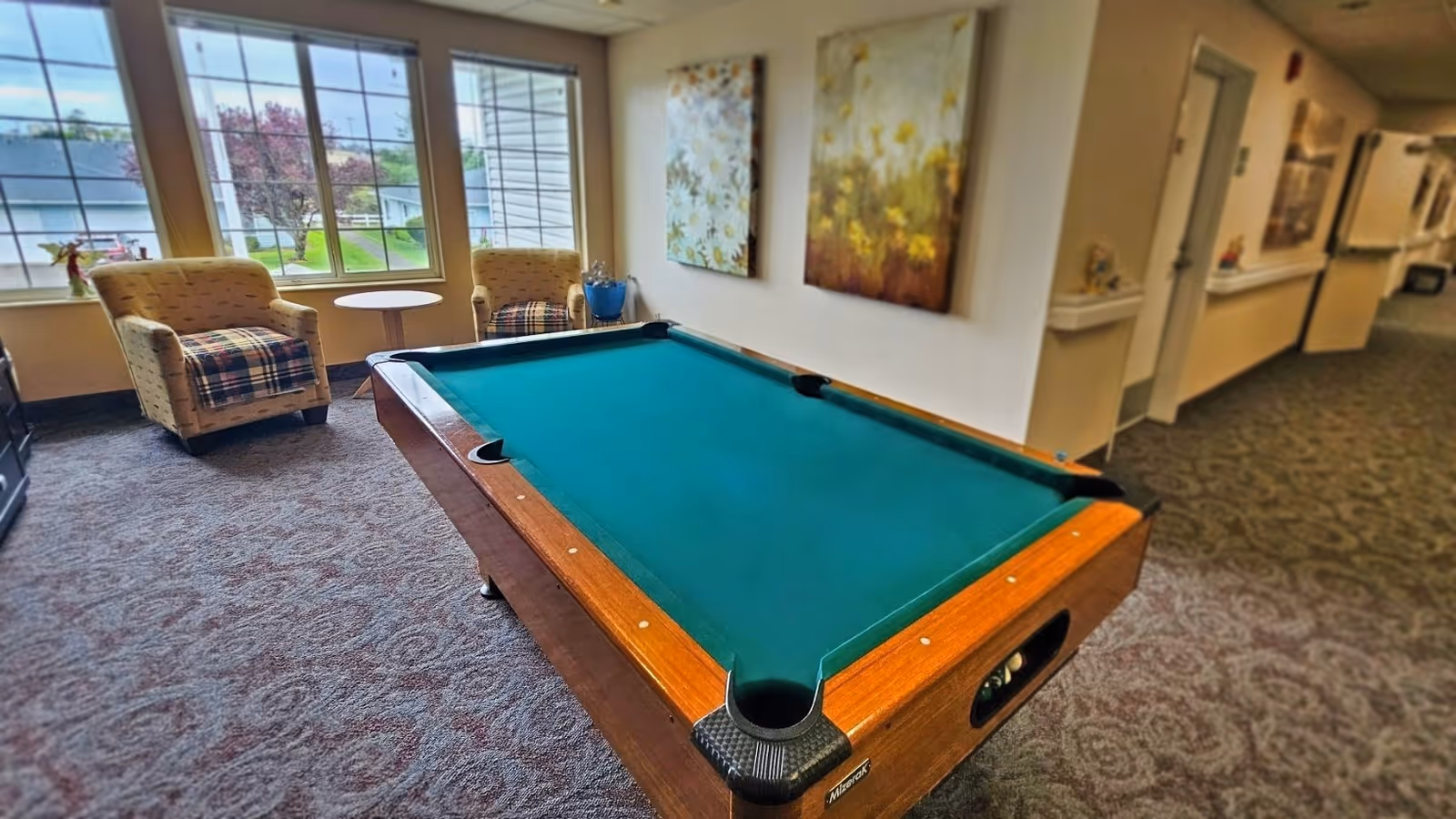 Interior view of a common area in a senior living facility featuring a pool table with a green felt surface. Two upholstered armchairs with plaid blankets are positioned near large windows letting in natural light. The walls are decorated with floral artwork, and a hallway extends to the right.