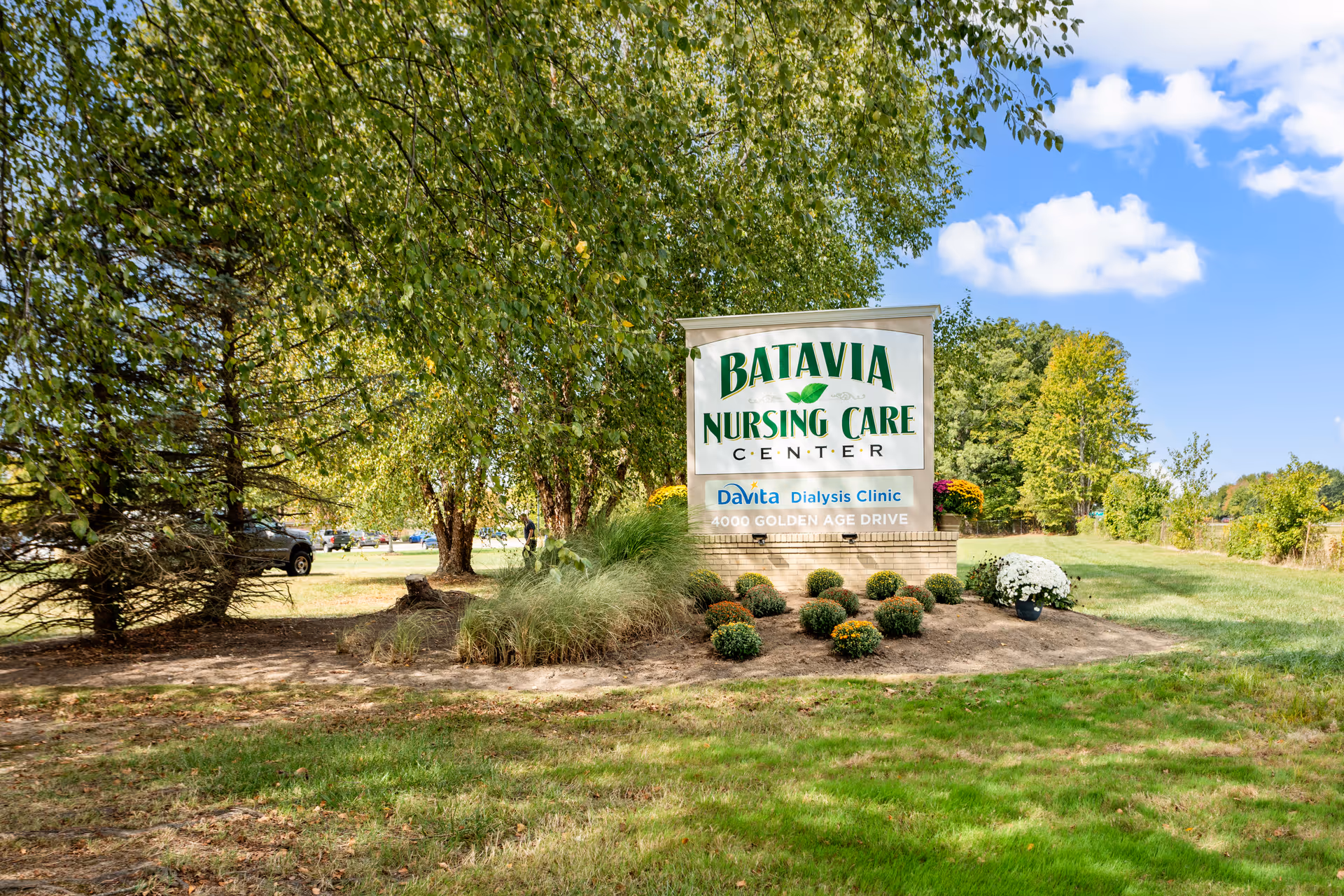 Outdoor view of the Batavia Nursing Care Center sign surrounded by landscaped bushes and trees under a blue sky with scattered clouds.