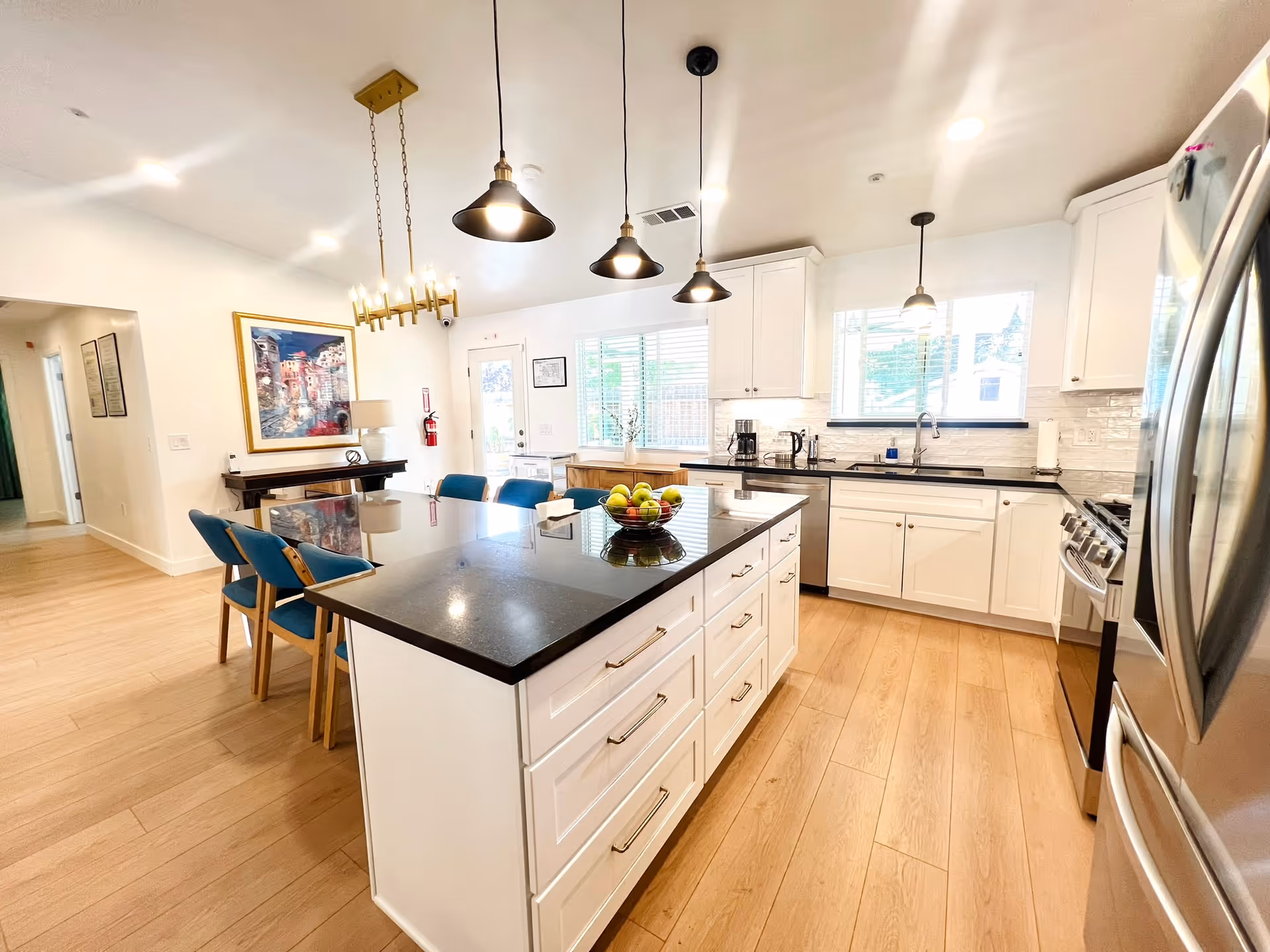 Bright and modern kitchen with white cabinets, black countertops, and stainless steel appliances. A large island with drawers and a bowl of fruit is in the center, surrounded by blue chairs. Pendant lights hang above the island, and a dining area with a chandelier and a framed painting is visible in the background.
