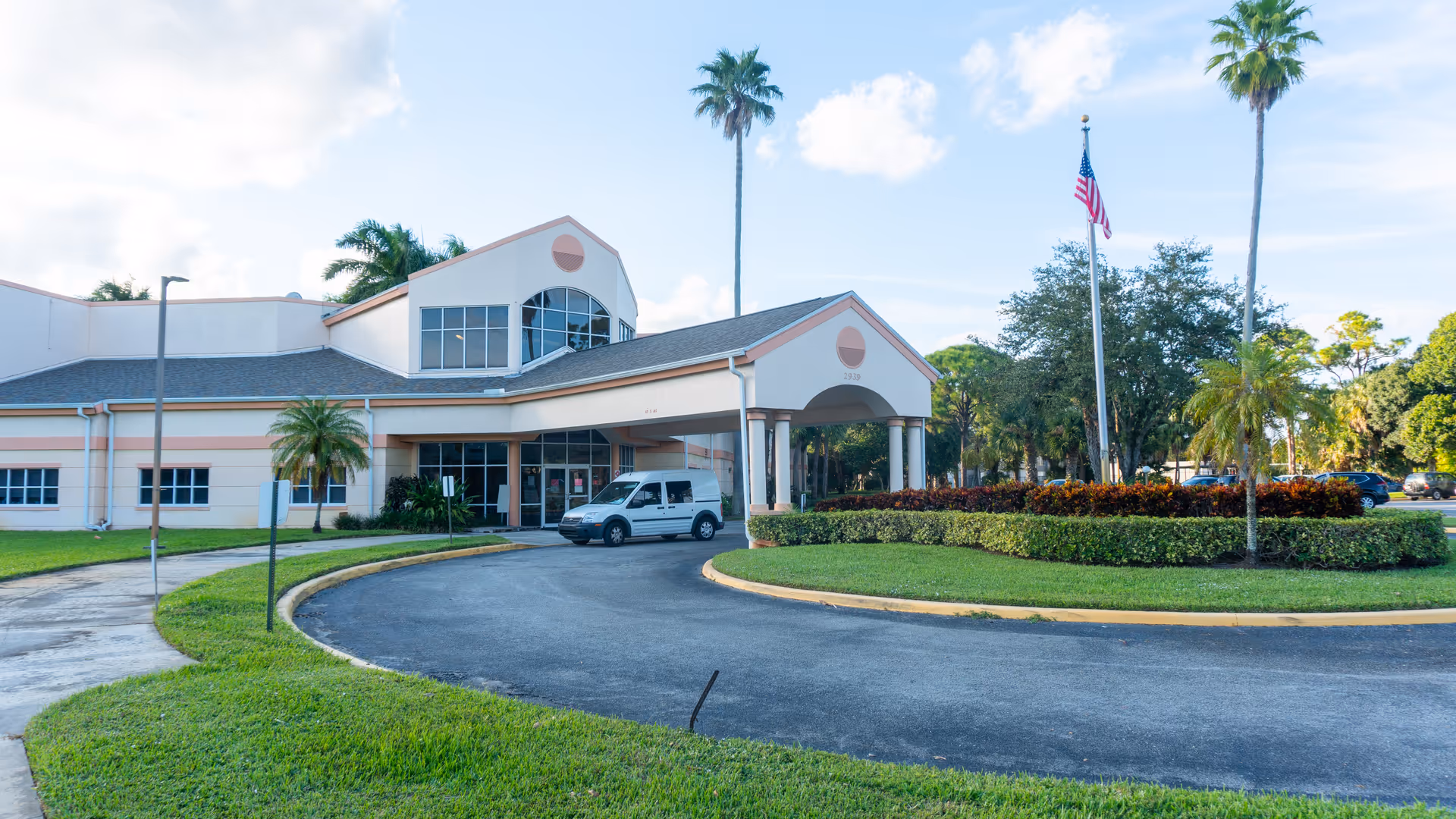 Exterior view of a senior living facility building with a covered entrance, a white van parked near the entrance, palm trees, a flagpole with an American flag, and well-maintained landscaping including grass and bushes.