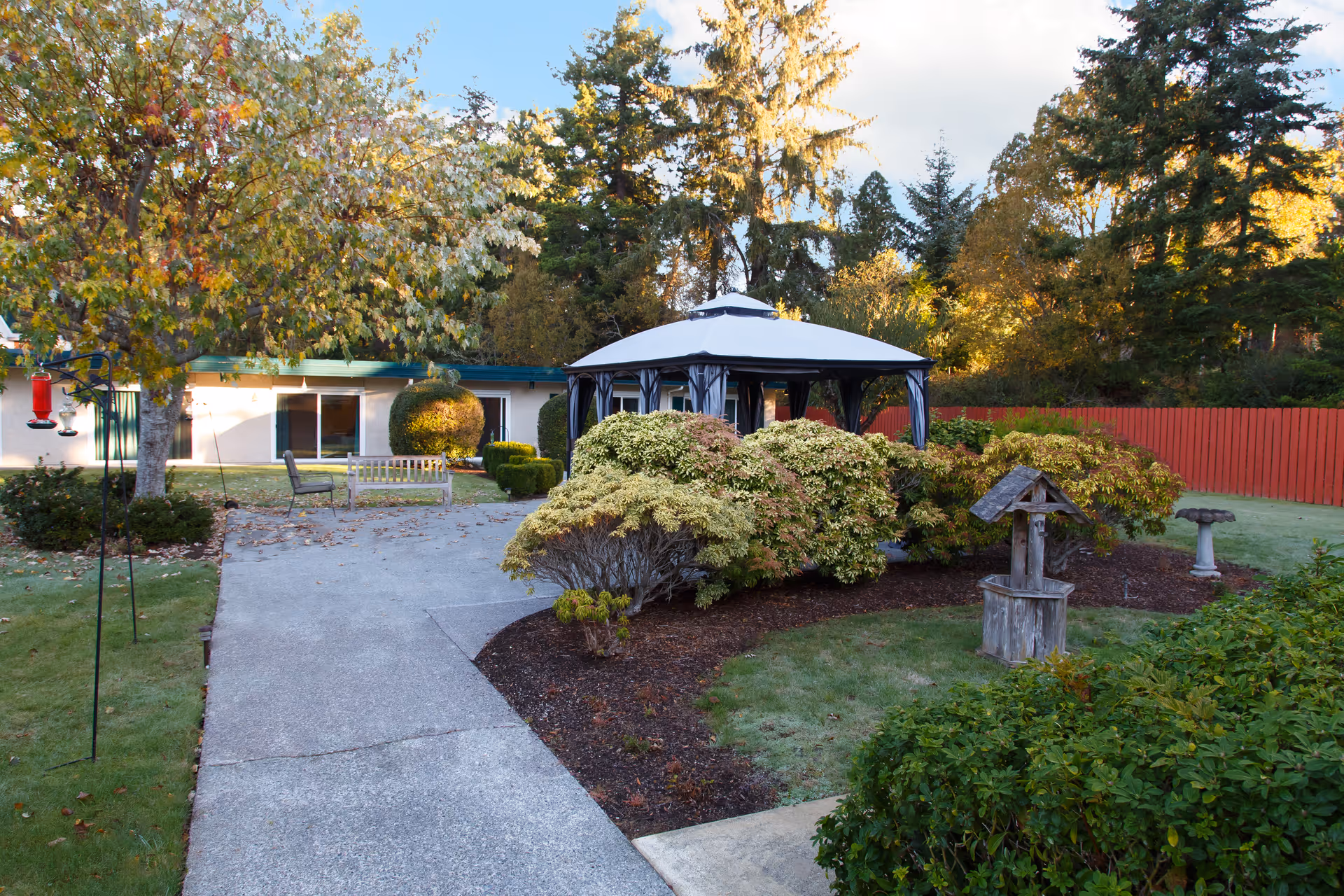 A landscaped courtyard with a paved path leading to a gazebo surrounded by shrubs, benches, and a low building in the background.