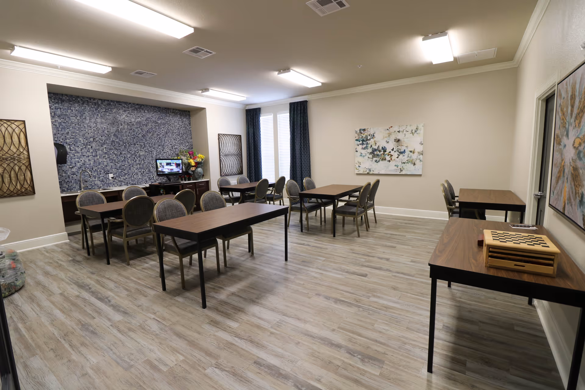 A well-lit room with several tables and chairs arranged for group seating. The room has light-colored walls and wood-style flooring. There is a small counter area with a mosaic tile backsplash, a computer monitor, and a vase with flowers. Two large windows with dark curtains allow natural light into the room. Artwork is displayed on the walls, and a chess/checkers board is placed on one of the tables.