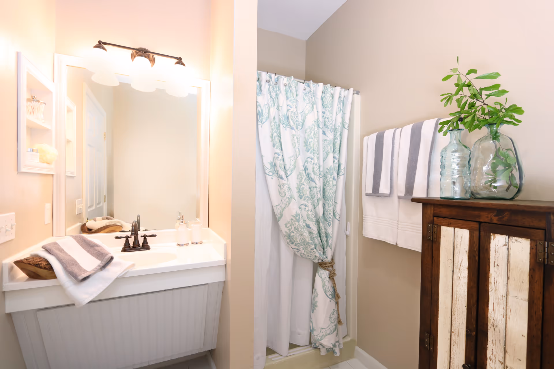 A bright bathroom featuring a white sink with a bronze faucet, a large mirror with three light fixtures above it, a shower with a white and green patterned curtain tied back with a rope, a wooden cabinet with distressed white panels, and two glass vases with green leafy branches on top. Two white and gray striped towels hang on a towel rack above the cabinet.