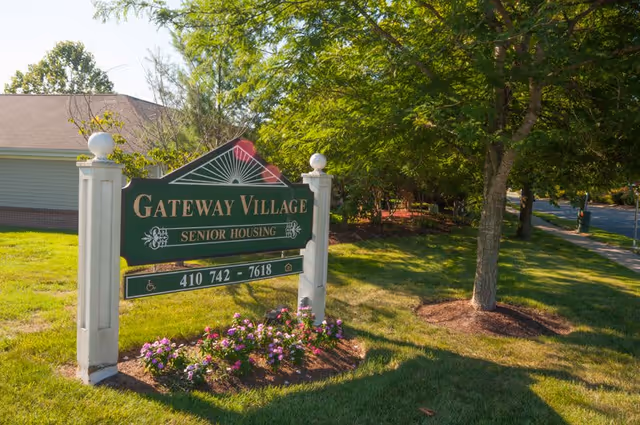 A green and white sign for Gateway Village Senior Housing is displayed on a grassy area with flowers planted at its base. The sign includes a phone number and accessibility symbols. Behind the sign, there is a building with a sloped roof and trees providing shade along a sidewalk and street.