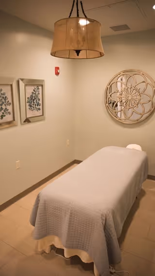 A small room with a massage table covered in a light gray quilted blanket. The walls are light-colored and decorated with two framed botanical prints on the left and a large round decorative mirror on the right. A beige pendant light hangs from the ceiling.