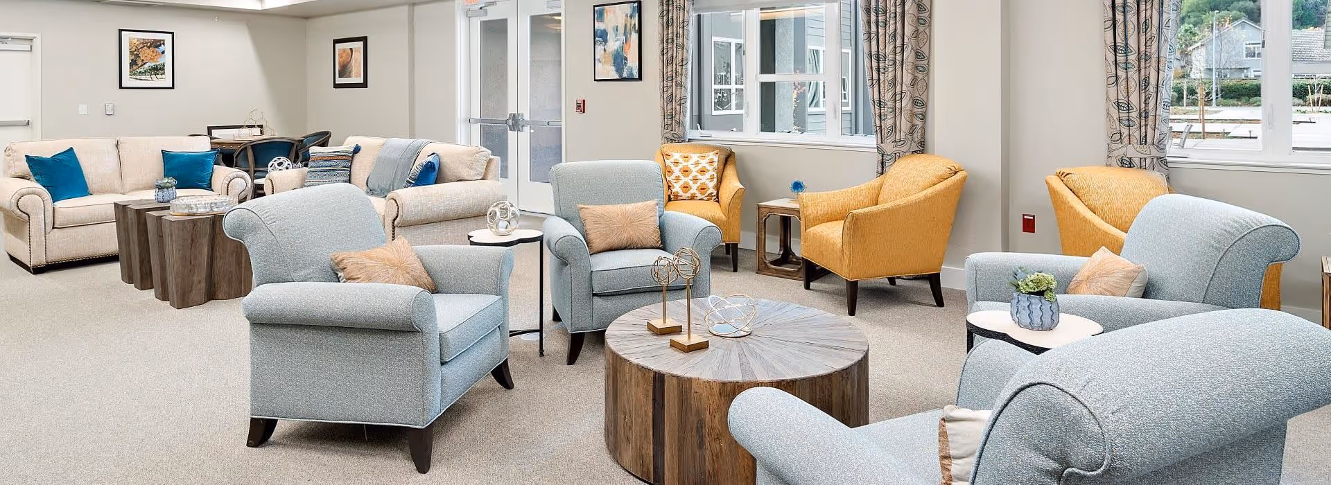 A bright and spacious living room area with multiple comfortable armchairs and sofas in soft blue and beige tones, accented with colorful throw pillows. There are round wooden coffee tables and side tables with decorative items. The room has large windows with patterned curtains allowing natural light to fill the space. Artwork is hung on the walls, and a dining table with chairs is visible in the background.