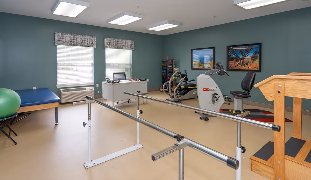 A physical therapy or rehabilitation room with parallel bars for walking exercises, an exercise bike, a recumbent bike, a wooden staircase for step training, a blue padded therapy table, a green exercise ball, a white desk with a chair, and two framed landscape pictures on the teal-colored walls.