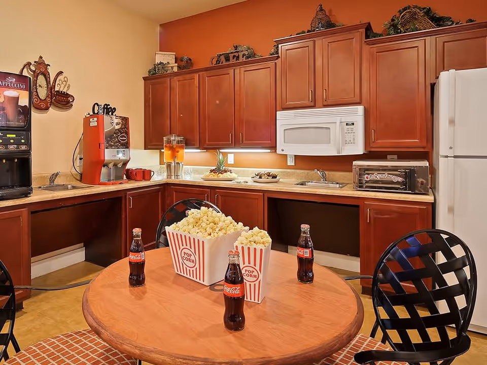 Small kitchenette with wooden cabinets, microwave, refrigerator, and a round table topped with popcorn boxes and Coca-Cola bottles.