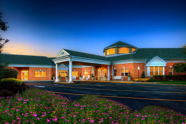 Exterior view of a single-story brick building with a covered entrance supported by white columns, illuminated windows, and a landscaped area with pink flowers in the foreground during twilight.