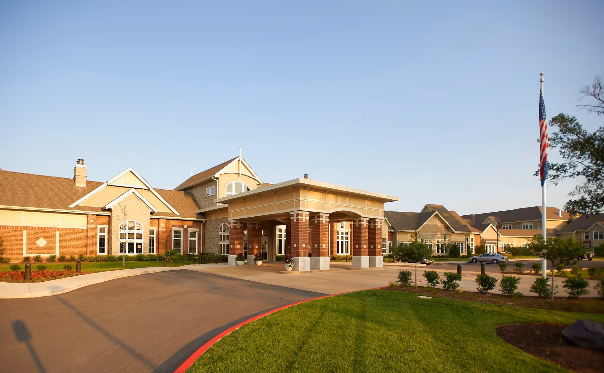 Front exterior of a large senior living facility with a covered porte-cochere entrance, landscaped grounds, and an American flag.