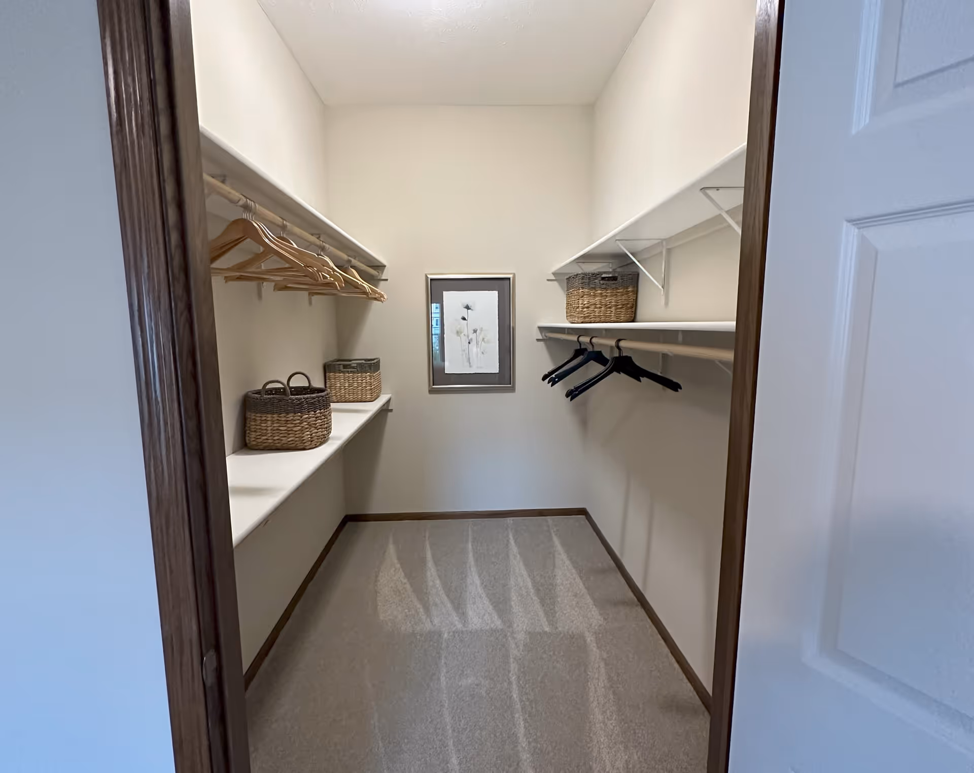 A walk-in closet with beige walls and carpeted floor. There are wooden and black hangers on white hanging rods on both sides. Two woven baskets are placed on a white shelf on the left side, and one woven basket is on a shelf on the right side. A framed floral artwork is hung on the back wall.
