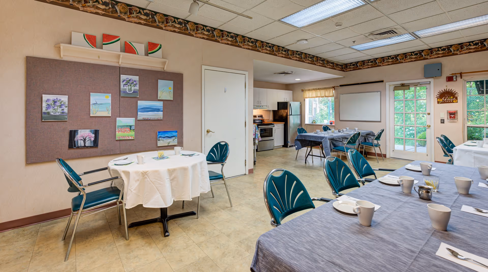 A dining area in a senior living facility with several tables covered in white and gray tablecloths. Each table is set with white cups, plates, and utensils. The room has teal chairs with metal frames. On the left wall, there is a bulletin board displaying various small paintings. In the background, there is a small kitchen area with a refrigerator, stove, and cabinets. A door with glass panels leads outside, and a window with a yellow valance lets in natural light.