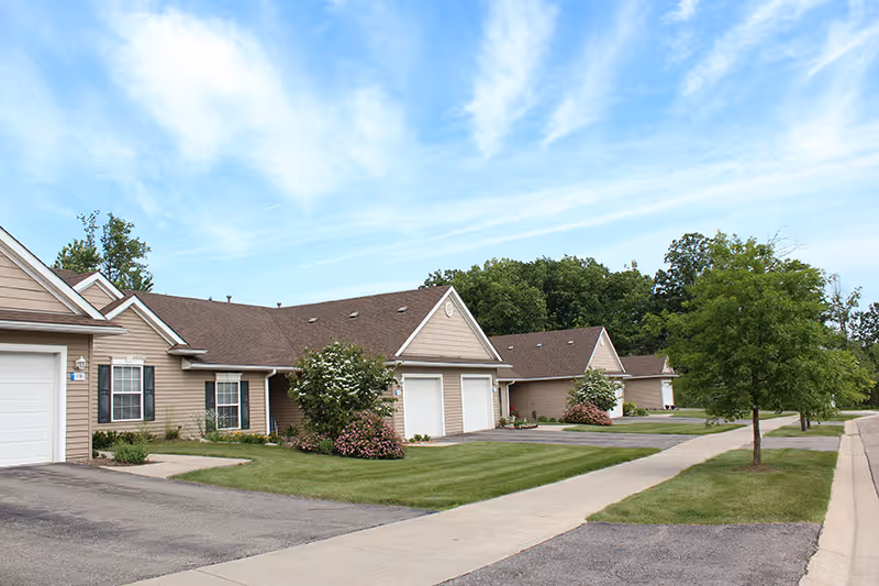A row of single-story beige townhome-style units with garages, driveways, manicured lawns, and a sidewalk under a blue sky.