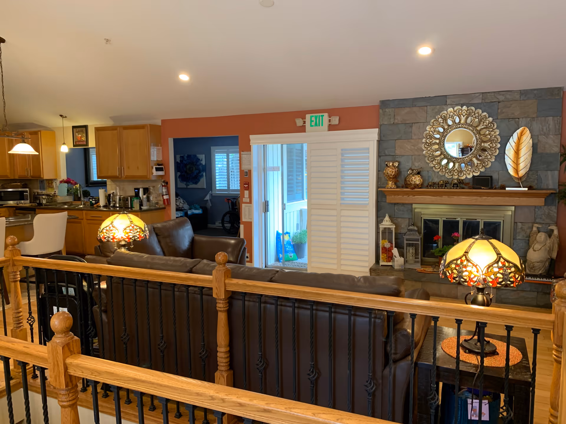 Interior view of a cozy living room with brown leather sofas, a stone fireplace adorned with decorative items including a round mirror and sculptures, and two stained glass table lamps. The room opens into a kitchen area with wooden cabinets and a dining space with a white chair. There is a sliding door with white shutters leading to an outdoor area, and a wooden railing with black metal spindles in the foreground.
