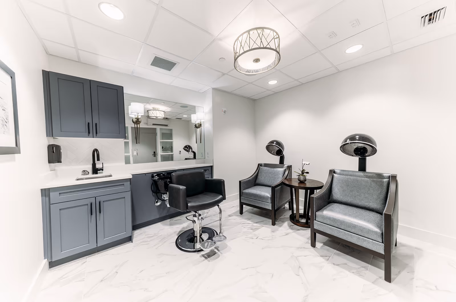 Bright salon-style interior with a styling station, sink and mirror on the left and two armchairs with hooded hairdryers on the right.
