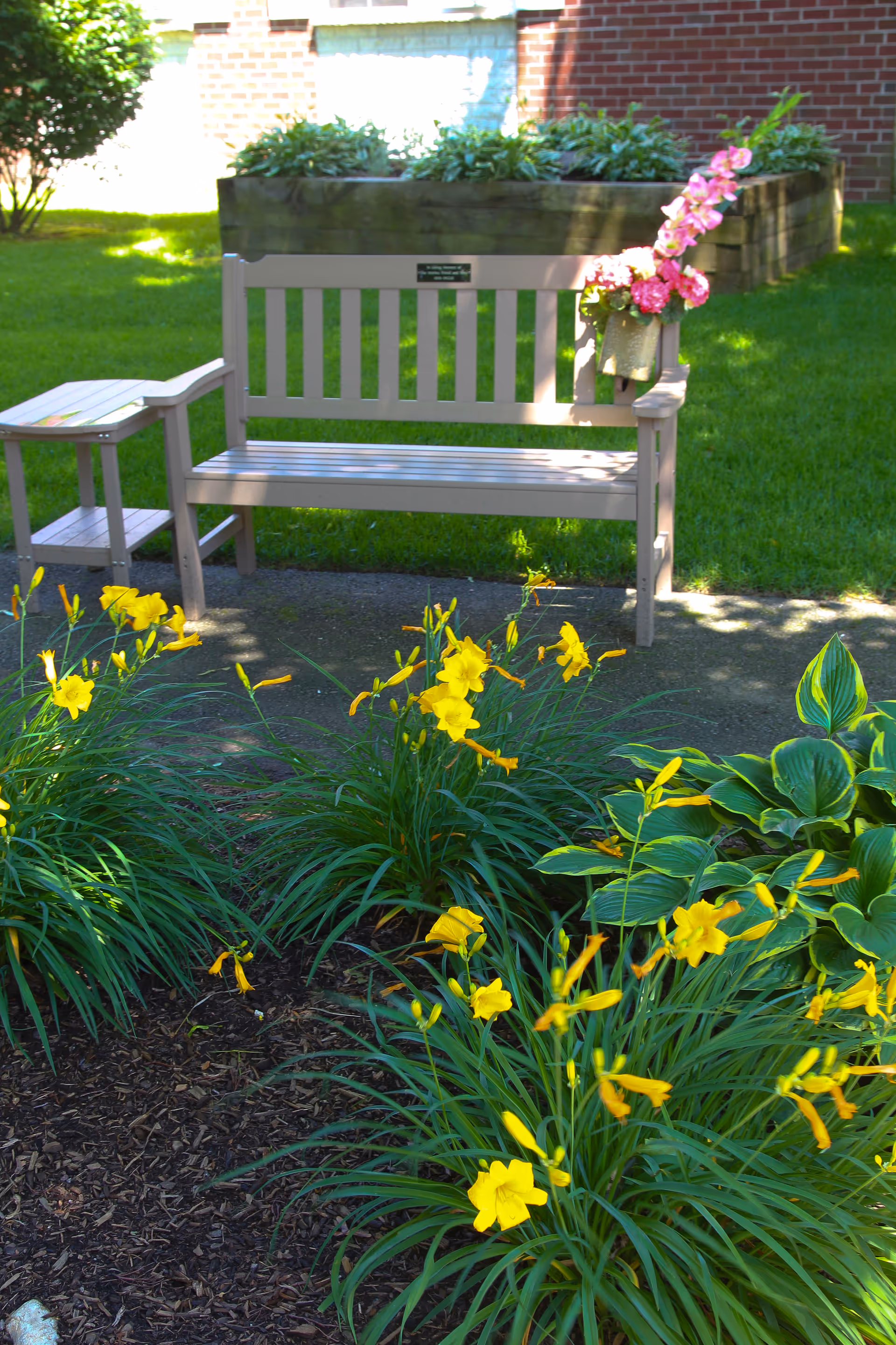 A wooden bench with a small side table attached, placed on a paved area in a garden. The bench has a small plaque and a flower pot with pink flowers hanging on one side. In front of the bench, there are clusters of yellow flowers and green plants. In the background, there is a raised garden bed and a brick wall.
