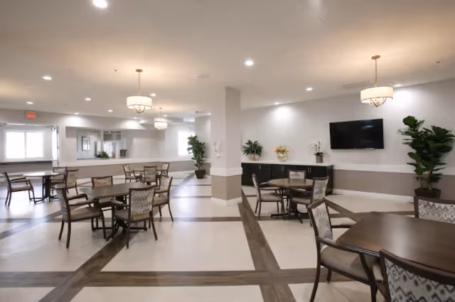 A spacious, well-lit common area with multiple tables and chairs arranged for seating. The room features light-colored walls, a patterned floor with dark wood accents, two chandeliers, potted plants, a wall-mounted flat-screen TV, and a sideboard with decorative items.