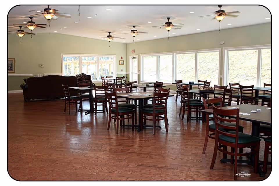 A spacious dining area with multiple wooden tables and chairs arranged neatly on a polished wooden floor. Large windows along one wall allow natural light to fill the room. Ceiling fans with lights hang from the white ceiling. There is a seating area with sofas in the background near the windows.