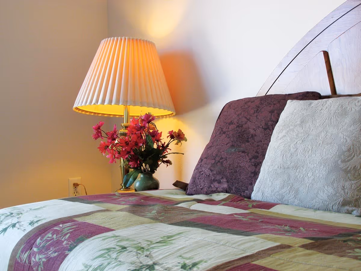 A cozy bedroom scene featuring a wooden headboard, two pillows with textured covers in purple and cream, a floral-patterned quilt, a brass table lamp with a pleated shade emitting warm light, and a small vase with pink flowers on a bedside table.