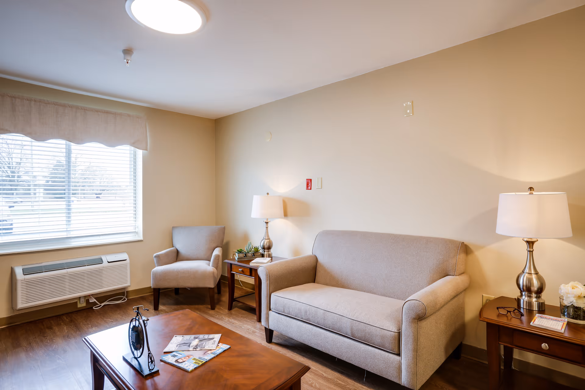A cozy living room with a beige sofa and matching armchair, two wooden side tables each with a lamp, a wooden coffee table with magazines and a decorative clock, a large window with blinds and a beige valance, and a wall-mounted air conditioning unit below the window.