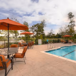 Outdoor pool area with several orange cushioned chairs and tables under large orange umbrellas, surrounded by a black metal fence and trees in the background under a partly cloudy sky.