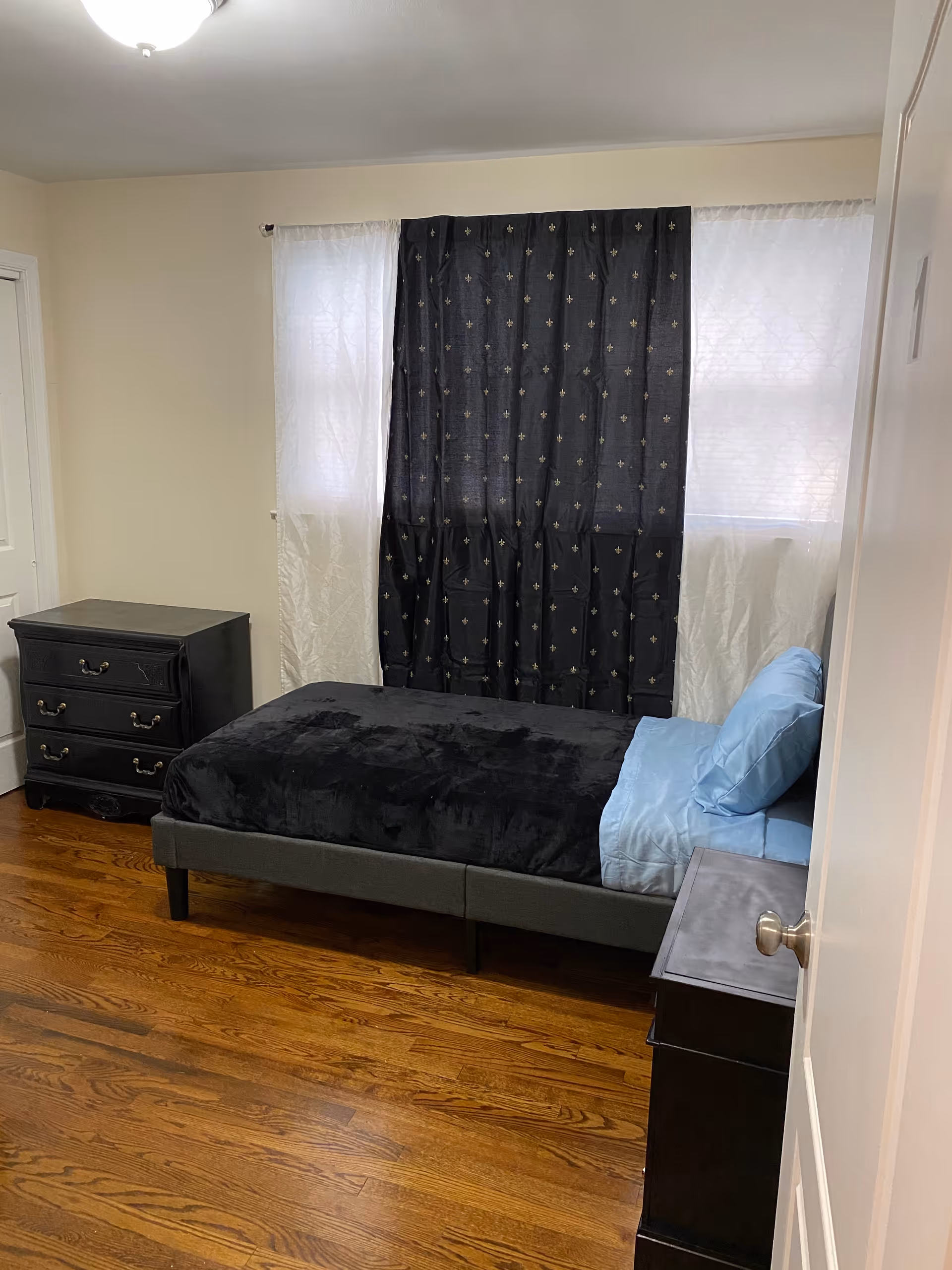Small bedroom with a single bed made with black bedding, a dresser and nightstands, and windows covered by dark curtains.