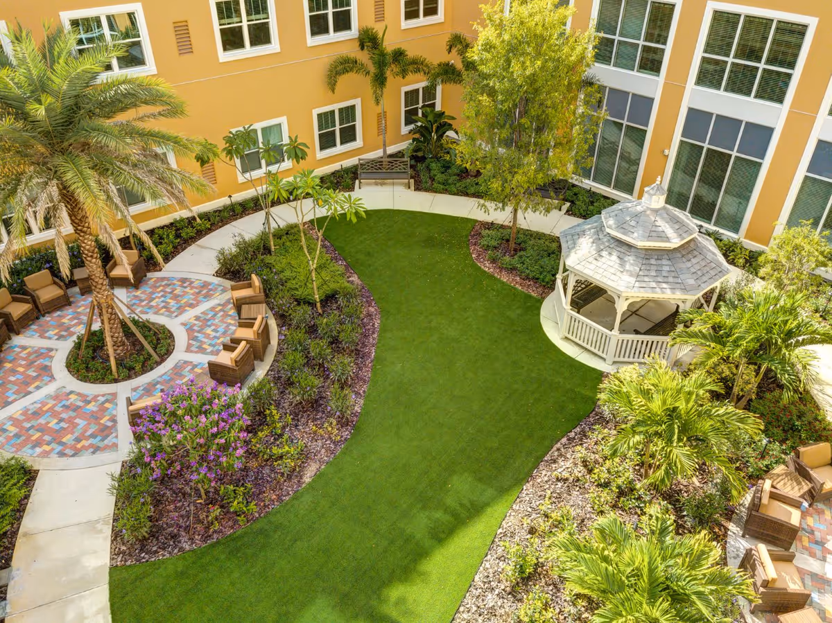 A landscaped outdoor courtyard area with a green lawn, palm trees, flowering plants, and a white gazebo. There are several cushioned chairs arranged on a circular patterned brick patio. The courtyard is surrounded by a yellow building with multiple windows.