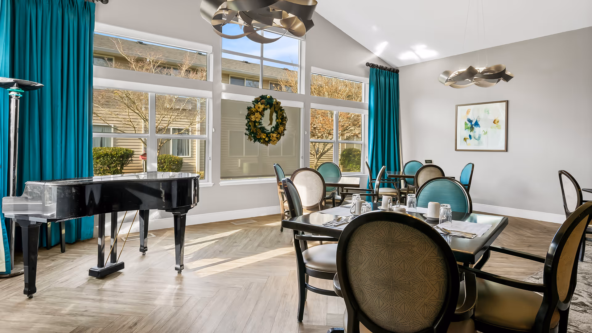 A bright dining room with large windows letting in natural light, teal curtains, a black grand piano, and several tables with chairs set for dining. A decorative wreath hangs on one of the windows, and a modern light fixture is suspended from the ceiling.