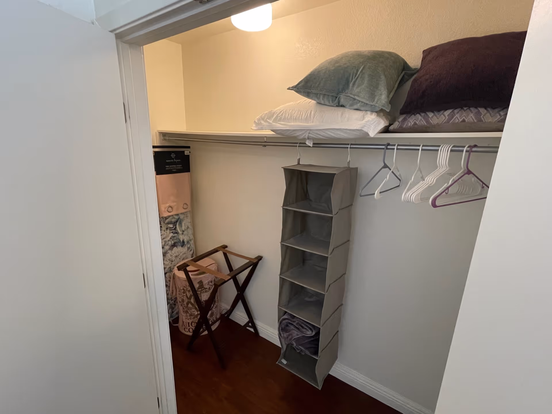 Interior view of a closet with a hanging organizer, several empty hangers, pillows on the top shelf, a folded blanket on the floor, and a wooden luggage rack.
