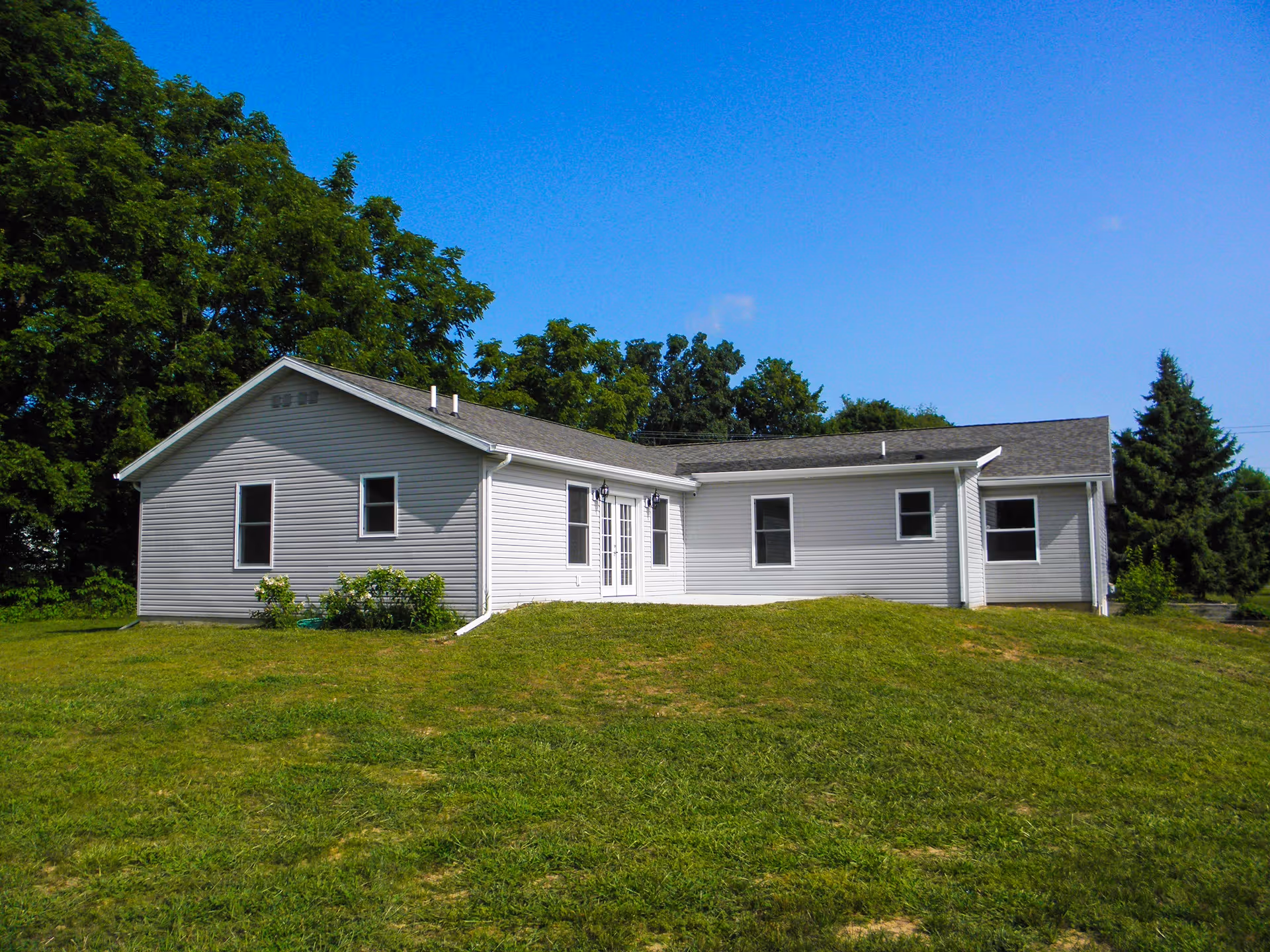 Single-story white building with multiple windows and a double door entrance, surrounded by green grass and trees under a clear blue sky.