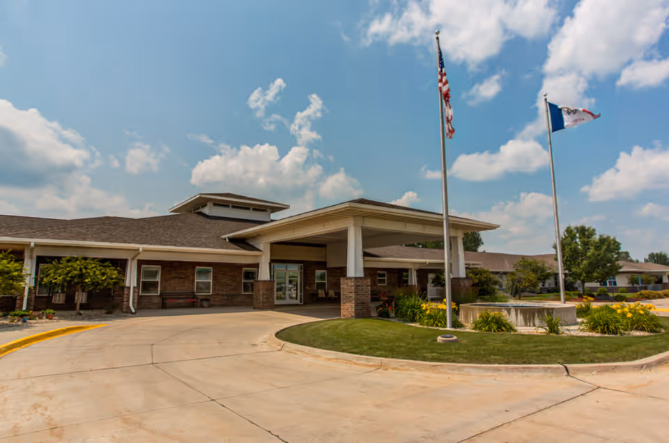 Exterior view of Prairie Hills at Tipton facility showing a single-story brick building with a covered entrance, two flagpoles with American and Iowa state flags, a circular driveway, and landscaped greenery under a partly cloudy sky.