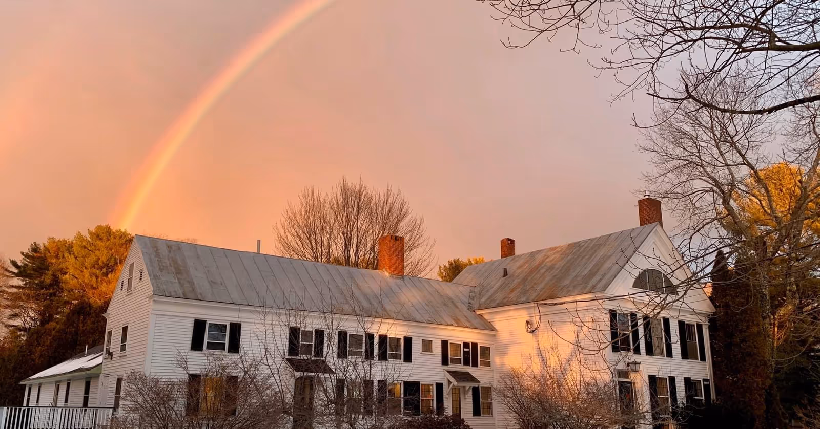 A large white building with black shutters and multiple chimneys under a sky with a visible rainbow and bare tree branches in the foreground.