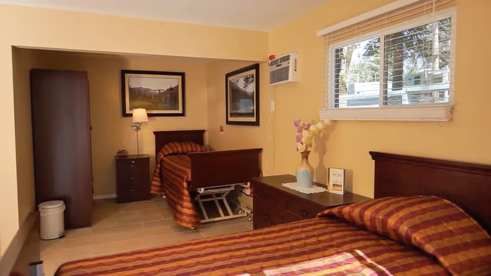 A bedroom in a senior living facility with two single beds featuring striped bedspreads in shades of red and orange. The room has light yellow walls, a window with white blinds, a wooden dresser with a vase of flowers and a small framed sign, a nightstand with a lamp, and framed artwork on the walls. There is also an air conditioning unit mounted on the wall.
