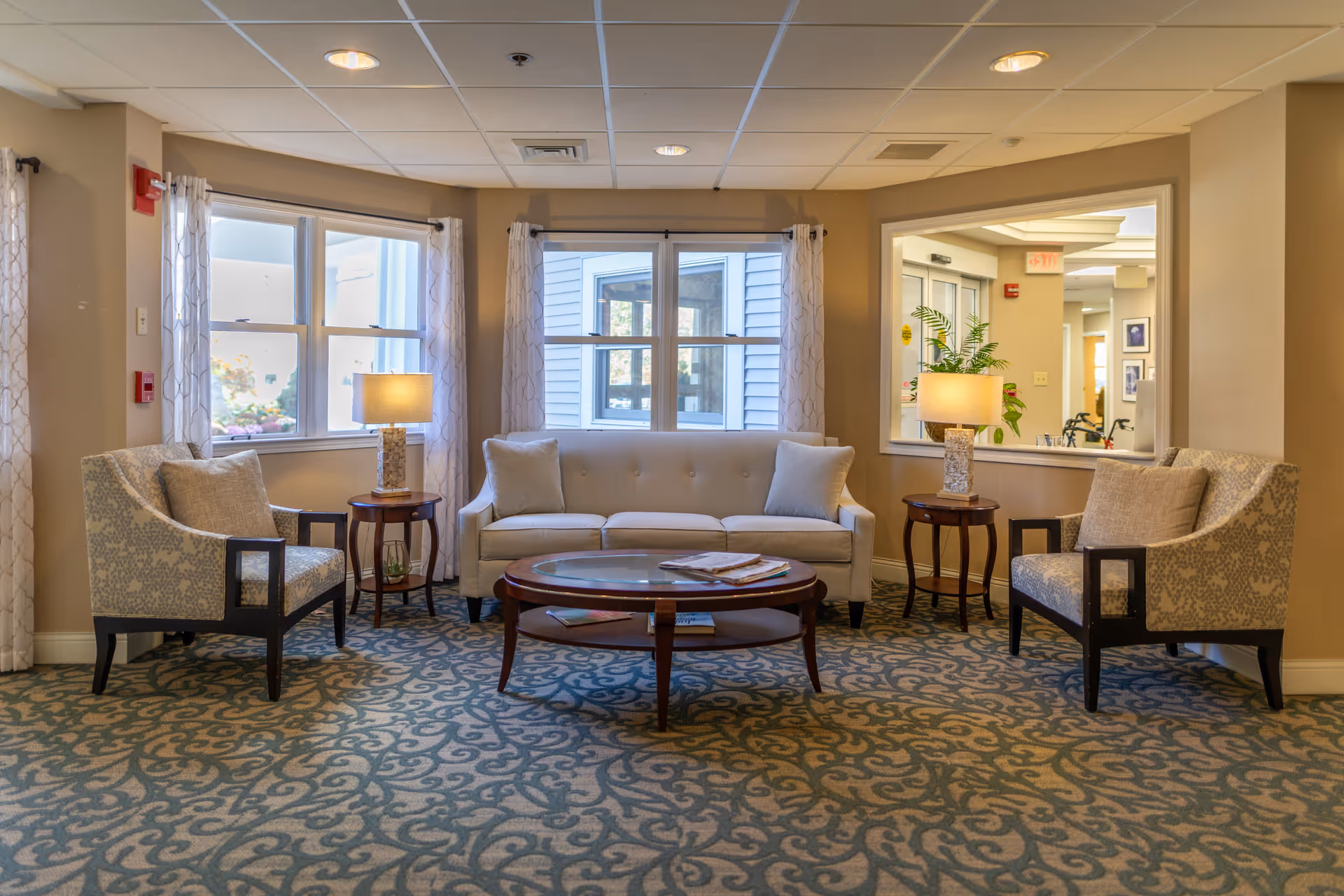 A cozy sitting area in a senior living facility with a beige sofa and two patterned armchairs arranged around a wooden coffee table. Two side tables with lamps flank the sofa. The room has large windows with sheer curtains, beige walls, and a patterned carpet. A window in the wall shows a hallway with plants and framed pictures.