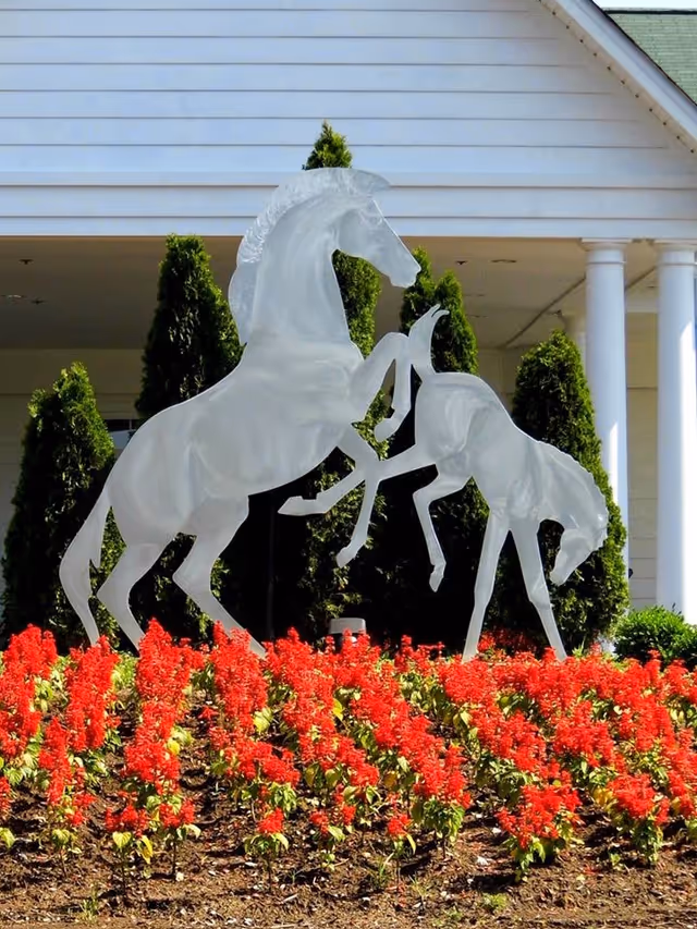 White horse sculptures, one rearing and the other bending down, placed in a garden bed with red flowers in front of a building with white siding and columns.