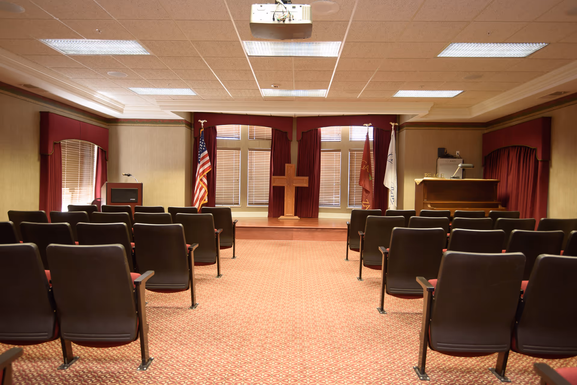 Interior view of a small chapel or meeting room with rows of chairs facing a stage with a wooden cross, American flag, and other flags. The room has red curtains, beige walls, and a patterned carpet.