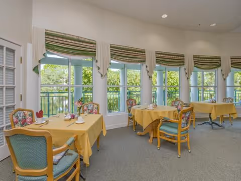 Bright dining room with small tables draped in yellow tablecloths and floral-upholstered chairs arranged along tall windows overlooking greenery and a pond.