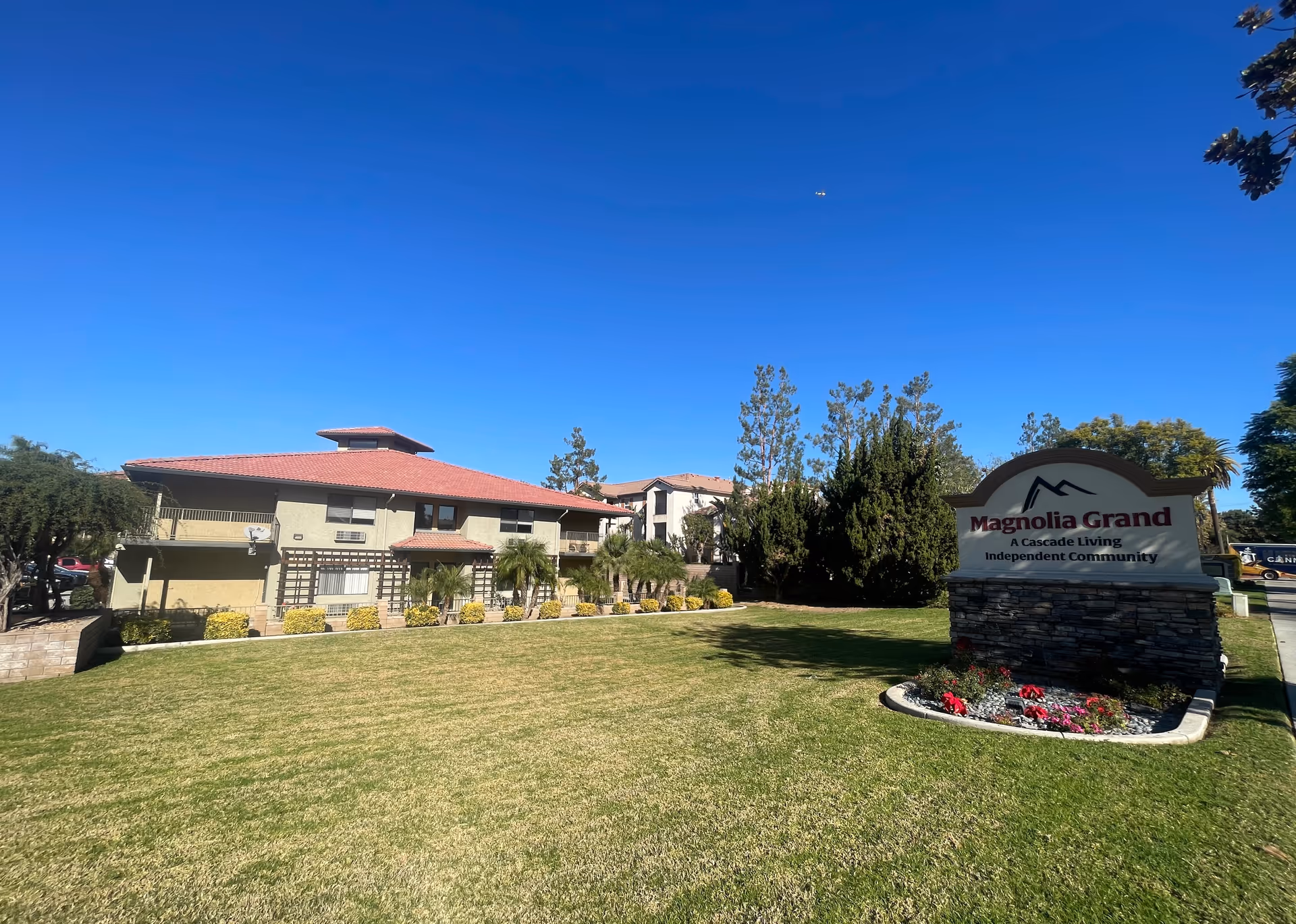 Exterior view of Magnolia Grand, an independent living community, showing a two-story building with a red-tiled roof, surrounded by trees and a well-maintained lawn under a clear blue sky. A stone sign with the community name and description is visible on the right side of the image.