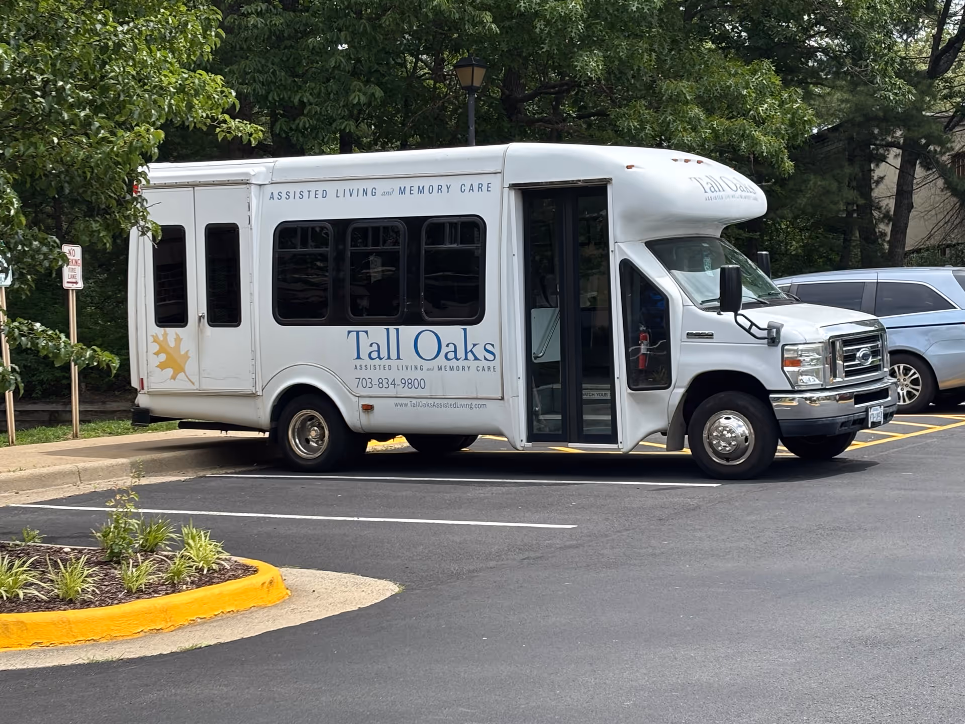 A white shuttle bus parked in a parking lot near some trees and other vehicles. The bus has signage for Tall Oaks Assisted Living and Memory Care, including a phone number and website. The bus door is open.