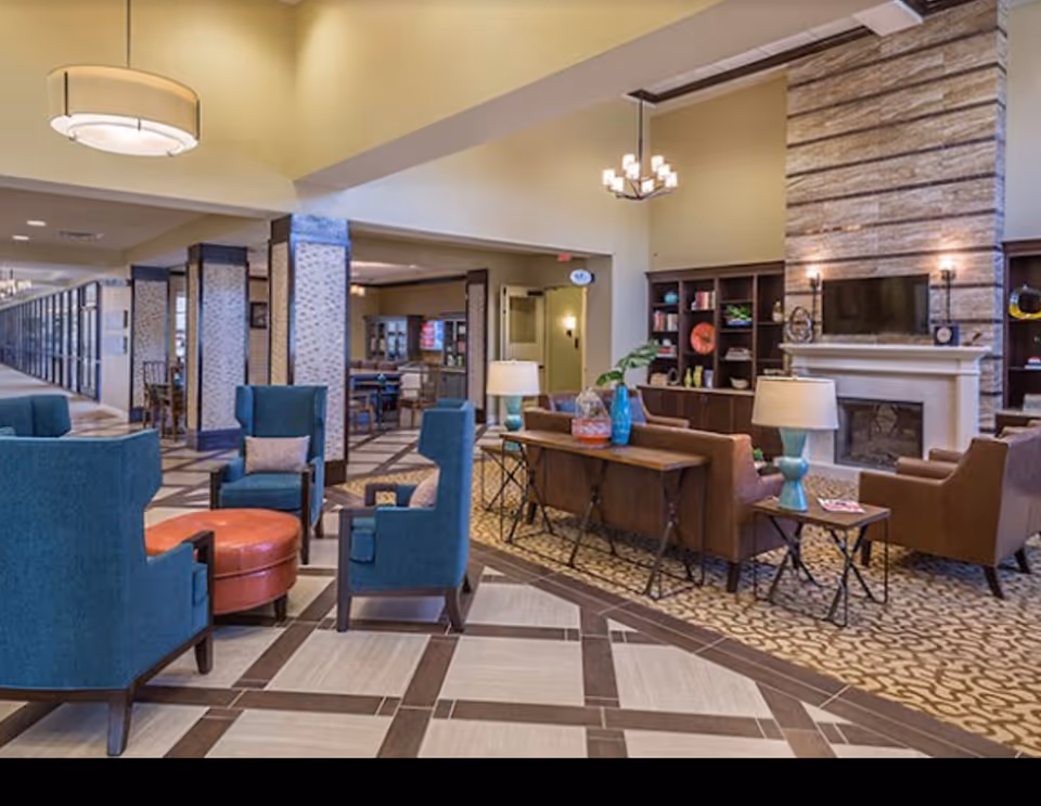 Spacious communal lounge with teal armchairs, brown sofas, a fireplace and built-in shelving under a high ceiling.
