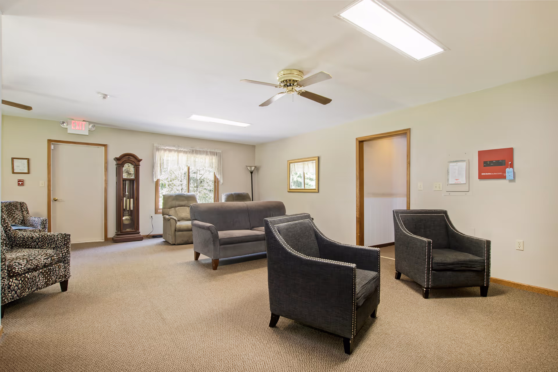A common area in an assisted living facility featuring a beige carpeted floor, two dark upholstered armchairs with nailhead trim, a gray loveseat, two recliners near a window with sheer curtains, a wooden grandfather clock, a ceiling fan, and a framed picture on the wall.