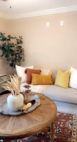 A cozy living room corner with a beige sofa adorned with white, rust, mustard, and yellow pillows. A wooden round coffee table in front holds a decorative tray with a white vase containing dried plants and other small decor items. A leafy green plant is placed in the corner near the sofa, and the walls are painted light beige with white crown molding. A patterned rug is partially visible under the coffee table.