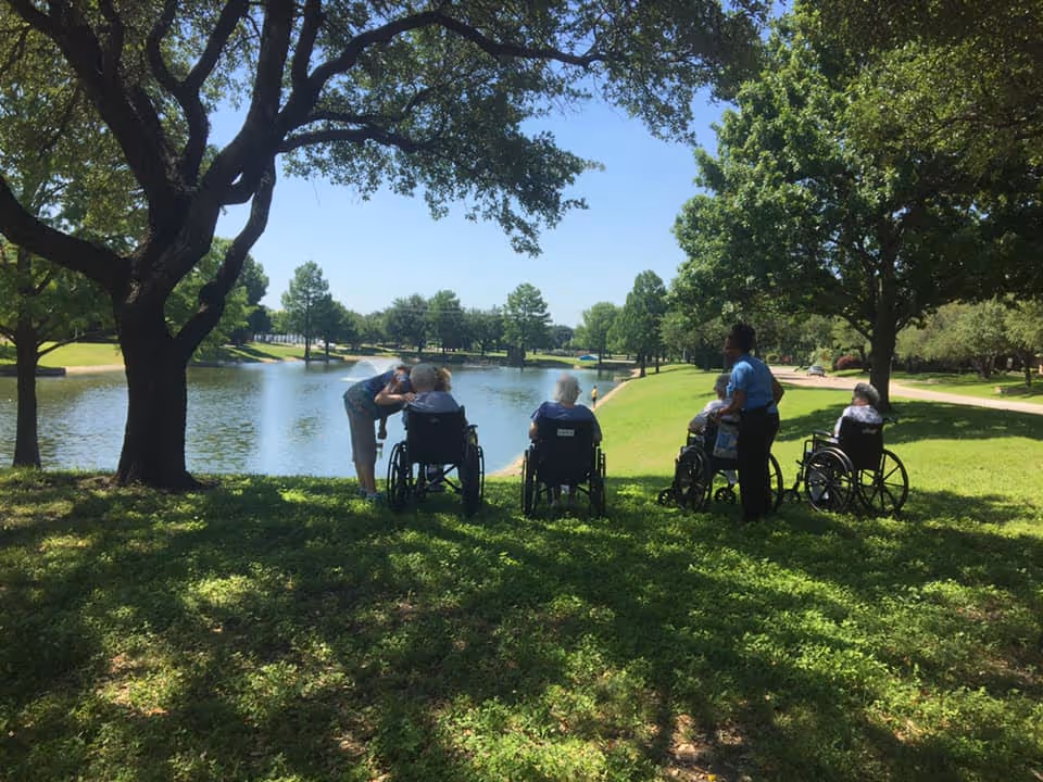 A group of elderly people in wheelchairs accompanied by caregivers are sitting and standing on a grassy area under large trees, overlooking a calm lake with a clear blue sky and more trees in the background.