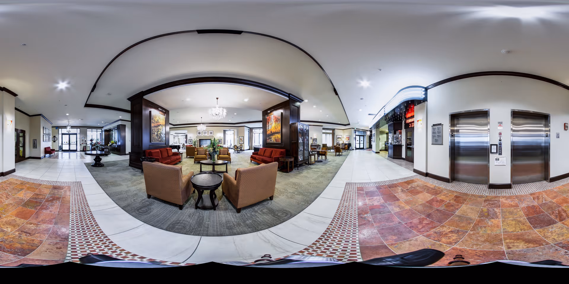 A spacious and well-lit senior living facility lobby with comfortable seating areas including armchairs and sofas arranged around small tables. The floor features a mix of tile and carpet, and there are two elevators on one side. The walls are decorated with large paintings and the ceiling has recessed lighting. The entrance doors are visible in the background.
